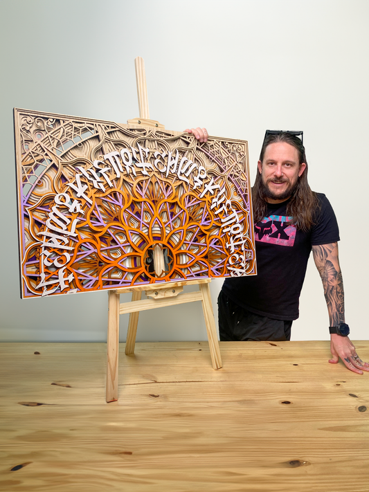 Artist Sketchy Brett standing beside his latest layered wooden artwork, Sunrise Strokes. The piece features intricate carved layers, blending natural wood tones with flowing, calligraphic details, capturing the warmth and movement of a sunrise.